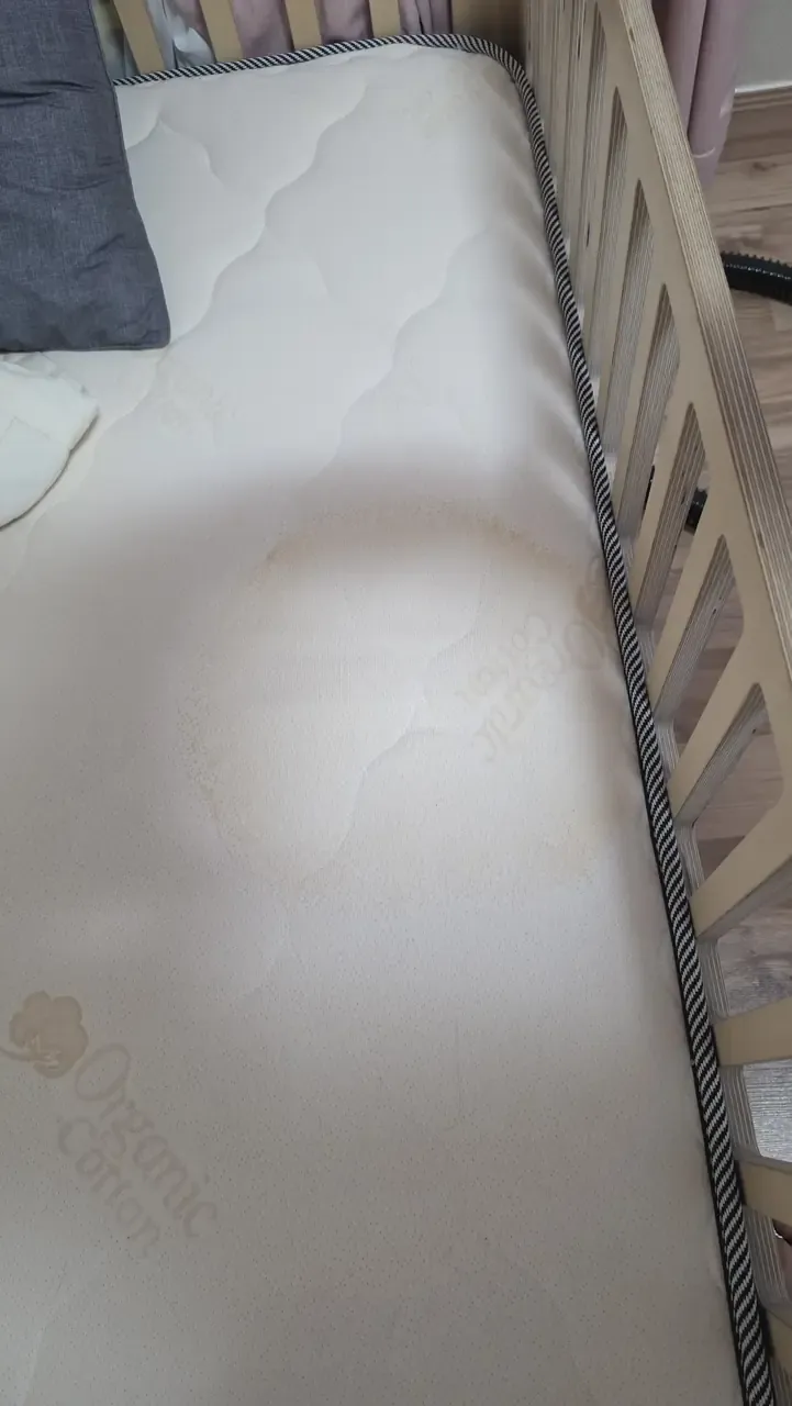 A cot mattress photographed from above with a large discoloured patch across the middle