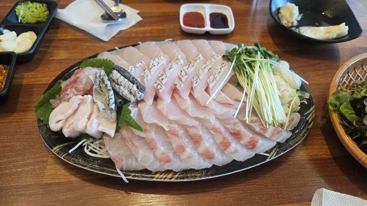 A large oval platter of thinly sliced white raw fish with perilla leaves, radish and dipping sauces on a wooden table in Seoul