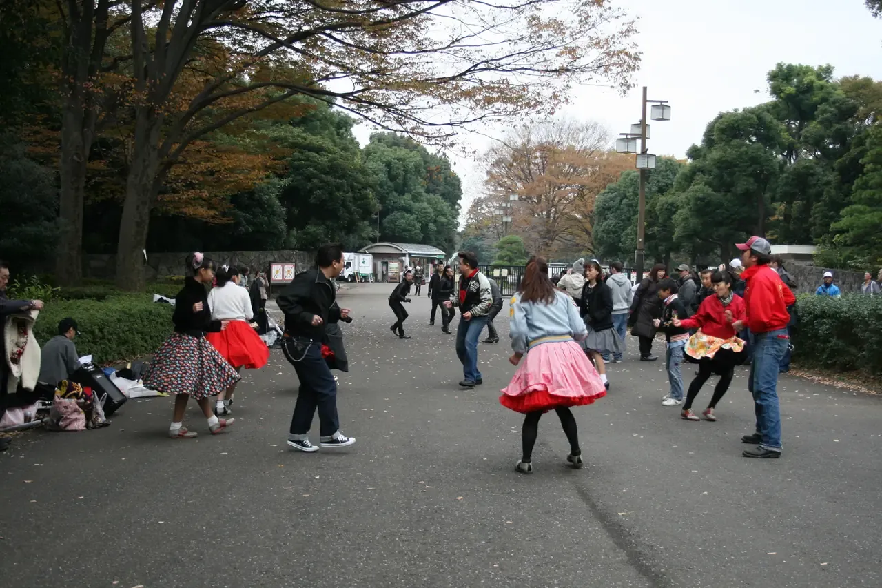 A group of people dancing outdoors in a park, in matching retro skirts and jackets, watched by passers-by